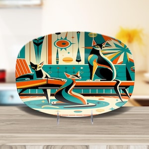 Op de afbeelding: Een ovale decoratieve schaal met een modern design uit het midden van de eeuw. Het toont drie gestileerde katten in een woonkamer. De katten zijn zwart, teal en oranje, tegen een teal en crème achtergrond.
