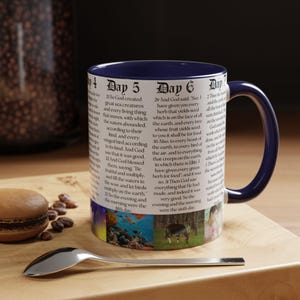 May include: A ceramic mug with a navy blue handle and interior. The mug features text from the Bible, with colorful images of sea life and animals. A spoon and a pastry are in the foreground.