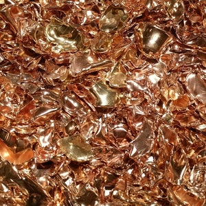 May include: A close-up of a pile of shiny, metallic copper flakes. The flakes are various sizes and shapes, and they have a warm, reddish-brown color.