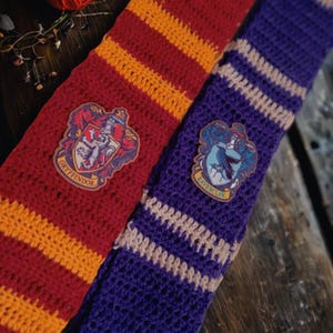 Peut inclure: Écharpes crochetées rouge et or, et violet et blanc, avec des écussons de maison. L'écharpe Gryffondor est rouge avec des rayures dorées, et l'écharpe Serdaigle est violette avec des rayures blanches. Présentées sur une surface en bois.
