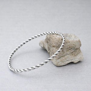 Peut inclure: Un bracelet jonc argenté avec un motif torsadé. Le bracelet repose sur une petite roche claire sur fond gris. Le bijou est un accessoire simple et élégant. Le diamètre du bracelet est d'environ 6,5 cm.