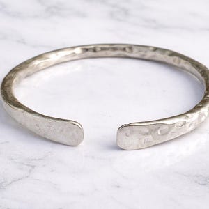 May include: A silver-coloured, open cuff bracelet with a hammered texture. The bracelet has a slightly irregular shape and is displayed on a white and grey marble surface. The ends of the bracelet are flattened.
