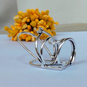 Op de afbeelding: Een zilverkleurige servetring met een uniek, open ontwerp. De ring heeft meerdere gebogen en geluste elementen, waardoor een abstracte vorm ontstaat. Op de achtergrond staat een tros gele bloemen.
