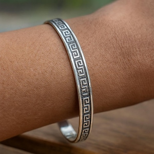 May include: A silver-colored bracelet with a Greek key pattern. The bracelet is worn on a wrist and has a polished finish. The design is a repeating geometric pattern. The bracelet is a piece of jewelry.