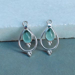 May include: A pair of silver teardrop earrings. Each earring has a light teal, teardrop-shaped stone set in a delicate frame. The design includes small, decorative silver accents at the top and bottom of the teardrop shape.