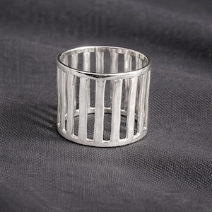 Könnte beinhalten: Ein silberner Ring mit einem käfigartigen Design. Der Ring hat vertikale Stäbe, die ein offenes, strukturiertes Aussehen erzeugen. Der Ring besteht aus glänzendem Metall und befindet sich vor einem dunkelgrauen Hintergrund.