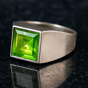 May include: A silver ring with a large, square-cut, bright green gemstone. The ring has a brushed metal finish and a thick band. The gemstone is a vibrant, eye-catching color, set in a square bezel.