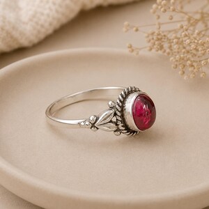 Pode incluir: Um anel de prata com uma grande pedra preciosa oval, vermelho escuro. O anel tem uma banda decorativa com detalhes de folhas e contas. A pedra preciosa é definida em uma moldura em forma de corda. O anel é exibido em um prato de tom neutro.