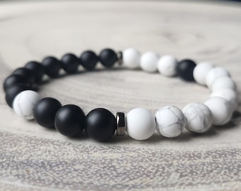 May include: A beaded bracelet featuring alternating black and white round beads. The bracelet has a mix of matte black onyx and white howlite beads, with a silver-colored spacer. The bracelet is a stylish accessory.