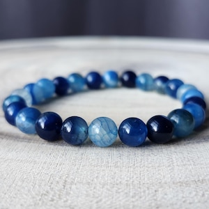 May include: A beaded bracelet featuring a variety of blue tones. The bracelet is composed of round, polished beads in shades of light blue, medium blue, and deep navy. The beads are strung together to form a complete circle.