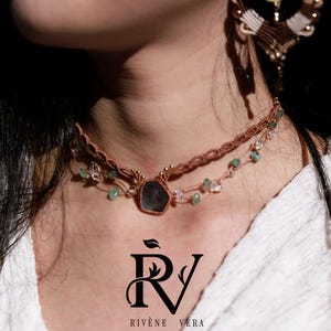 May include: A double-strand brown macrame choker necklace featuring a central dark stone, small green and clear beads, and a gold-tone crescent moon earring. The logo RIVÈNE VÉRA is printed in black below the necklace on the white fabric.