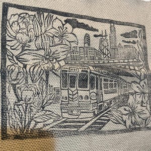 May include: A black and white illustration of a city scene on a beige fabric. The image features a trolley car, skyscrapers, and floral elements. The trolley car is on tracks, and the skyscrapers are in the background.
