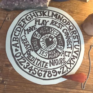 May include: A round, white divination board with black lettering and a heart design. The board features letters, numbers, and words like "Yes," "No," and "Ask Again." A red, teardrop-shaped planchette rests on the board, with a silver chain attached.