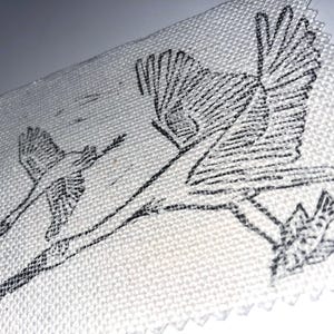 以下が含まれることがあります： 白い布のクローズアップ。黒インクで鳥と枝が描かれています。デザインは、飛んでいる鳥と枝に止まっている鳥を特徴としています。生地は質感のある外観をしています。