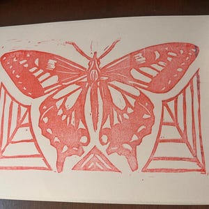 May include: A red linocut print of a butterfly with detailed wing patterns. The butterfly is centered on the paper, with geometric shapes below the wings. The print is on white paper.