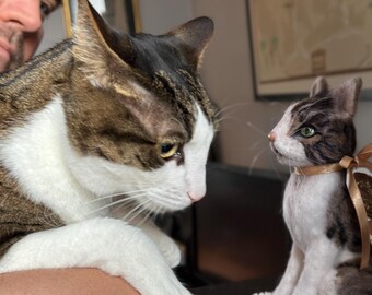 May include: A tabby cat with white patches gazes at a small, detailed cat figurine. The figurine has a brown and white pattern, green eyes, and a ribbon. The cats appear to be interacting, creating a charming scene.