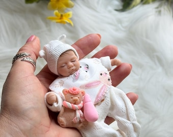 Può includere: Una bambola neonata in miniatura con cappello e tutina in maglia bianca, tenuta in mano. La bambola ha gli occhi chiusi e un'espressione serena. Un piccolo orsacchiotto con sciarpa rosa e un biberon rosa sono accanto alla bambola. Fiori gialli e pelliccia bianca sullo sfondo.