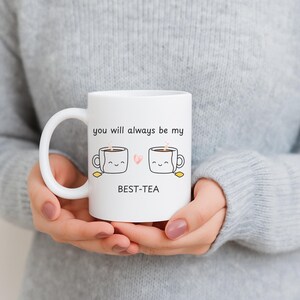 Könnte beinhalten: Weißer Keramikbecher mit gebogenem Henkel, mit dem Text "you will always be my BEST-TEA". Der Becher hat ein Design von zwei lächelnden Teetassen mit Dampf und einem rosa Herz dazwischen. Der Becher wird von einer Person gehalten, die einen grauen Pullover trägt.