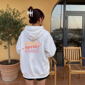 Könnte beinhalten: Weißer Kapuzenpullover mit dem Text "WOMEN SUPPORT WOMEN" in Orange und Rosa. Der Kapuzenpullover hat eine Kapuze mit Kordelzug und eine Kängurutasche. Das Bild zeigt die Rückseite des Kapuzenpullovers.