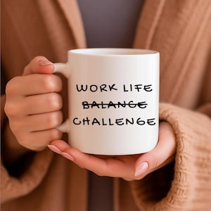Könnte beinhalten: Eine weiße Keramik-Tasse mit den Worten "WORK LIFE BALANCE CHALLENGE" in Schwarz gedruckt. Die Tasse wird von einer Person gehalten, die einen braunen Pullover trägt.