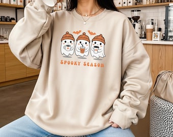 May include: Beige sweatshirt with a graphic of three cartoon ghosts wearing orange beanies and holding coffee cups. The text "SPOOKY SEASON" is printed in orange below the ghosts. The sweatshirt has long sleeves and a crew neck.