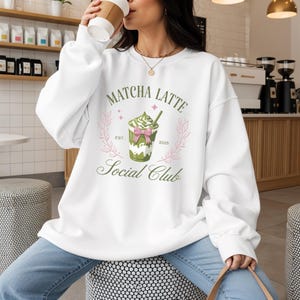 Könnte beinhalten: Weißes Sweatshirt mit einem Matcha Latte-Motiv. Das Design zeigt einen grünen Matcha Latte mit Schlagsahne, einer rosa Schleife und dem Text "Matcha Latte Social Club". Das Sweatshirt hat einen lässigen Stil.