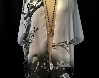 May include: A sheer, open-front kimono with a black and white floral and bird print. A long beaded necklace with a gold-colored Hamsa hand pendant hangs in front of the kimono. The kimono is displayed on a mannequin against a black background.