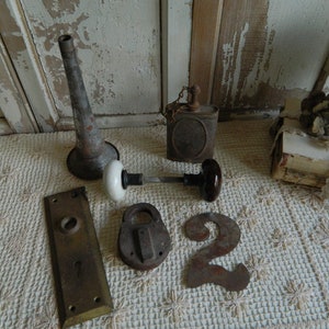 May include: An assortment of vintage metal objects, including a funnel, a small oil can, a door knob, a padlock, a number 2, and a door plate. The items are arranged on a cream-colored, textured surface.