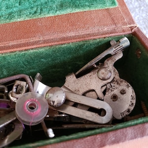 May include: A close-up of a vintage sewing machine foot pedal and other metal sewing machine parts in a green velvet-lined case. The parts are silver and have a worn, aged look.