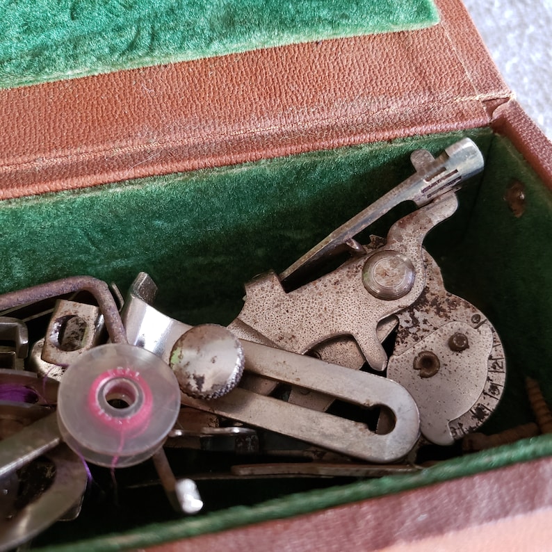 May include: A close-up of a vintage sewing machine foot pedal and other metal sewing machine parts in a green velvet-lined case. The parts are silver and have a worn, aged look.