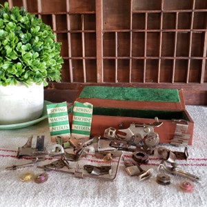 May include: An open brown box with green felt lining contains a variety of sewing machine parts. Two small packages of sewing machine needles are visible on the table next to the box. The packages are labeled "Sewing Machine Needles".