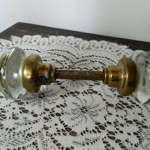 May include: Antique glass door knob with brass hardware. The clear glass knobs have a rounded shape, and the brass components show signs of age. The connecting rod is a rusty brown color. This vintage hardware adds a touch of classic charm.
