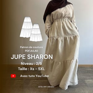 May include: A beige tiered maxi dress with long sleeves and a gathered waist. The image includes a pattern illustration and text that reads "JUPE SHARON" and "Avec tuto YouTube". The dress is shown on a person wearing a brown head covering.