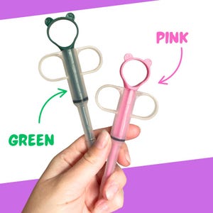 Puede incluir: Dos dispensadores de medicamentos para beb&eacute;s, uno verde y otro rosa, se sostienen en una mano. El dispensador verde tiene una parte superior en forma de oso y la palabra "GREEN" en verde. El dispensador rosa tiene una parte superior en forma de oso y la palabra "PINK" en rosa.