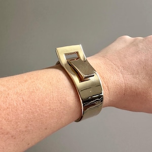 May include: A gold-colored cuff bracelet with a rectangular buckle design. The bracelet is shiny and appears to be made of metal. The buckle is a prominent feature, adding a modern touch to the jewelry.