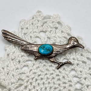 May include: A silver-toned metal roadrunner pin with a turquoise stone set in the center. The bird is depicted in a running pose, with detailed feather and leg features. The pin is set against a white, lacy background.