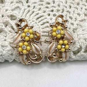 May include: A pair of vintage gold-tone earrings with yellow flower clusters and white enamel leaves. Each earring features a central clear rhinestone and small gold beads. The earrings are set against a white crocheted background.