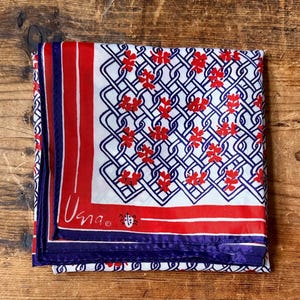 May include: Folded square scarf with a white background, featuring a red and blue geometric pattern and red floral accents. The scarf has a red and blue border and the word "Usna" is printed on the scarf.