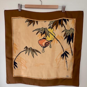 May include: A square silk scarf with a brown border and a central illustration. The illustration features bamboo stalks, two hanging lanterns in yellow and orange, and a bridge. The background is a light tan color.