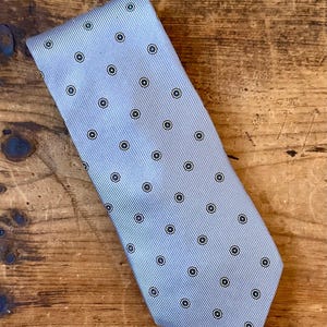 May include: A light blue tie with a repeating pattern of small black and white circles. The tie has a ribbed texture and is displayed on a wooden surface.