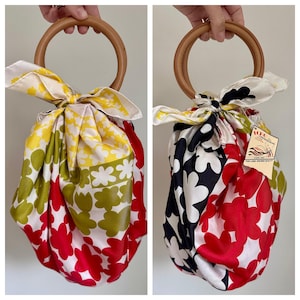 May include: A fabric pouch bag featuring a round wooden handle and a colorful floral pattern in red, yellow, green, and white. The bag is tied at the top with a silk-like scarf. A tag attached to the bag reads Echo Creations, Since 1923.
