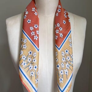 May include: A floral patterned scarf with a peach and yellow background. The scarf features white flowers with blue centers and blue and white stripes. The scarf is draped over a mannequin.
