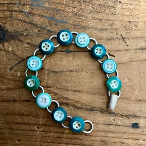 May include: A bracelet made of silver chain links with green and white buttons. The buttons have a white center with four holes and a green outer ring.