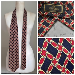 May include: A navy blue silk tie with a repeating pattern of red and gold chain links. The tie is displayed on a mannequin. The label reads "FENDI" and "Made in Italy".
