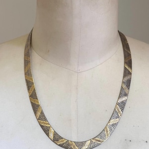 May include: A silver-toned necklace with a geometric pattern of gold and silver chevrons. The necklace is displayed on a mannequin and has a flat, wide design. The metal has a textured appearance, and the necklace is suitable for formal or casual wear.