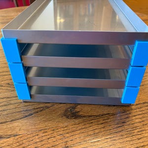 Könnte beinhalten: Ein silberner Metall-Organizer mit vier Regalen. Die Ecken sind mit leuchtend blauen Kunststoffteilen akzentuiert. Der Organizer steht auf einer Holzoberfläche.