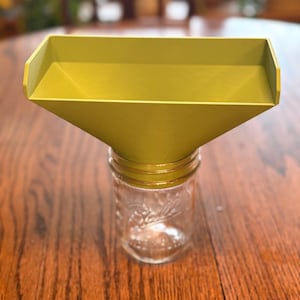 May include: A yellow-green funnel is placed over a clear glass jar. The funnel has a rectangular top and tapers down to a smaller opening. The jar has a decorative design. The items are on a wooden surface.