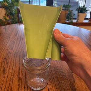 Peut inclure: Entonnoir en plastique vert clair au-dessus d'un bocal en verre transparent. L'entonnoir a un dessus évasé et large et un bec rectangulaire. Le bocal est vide et posé sur une table en bois. L'arrière-plan comprend une fenêtre et des plantes.