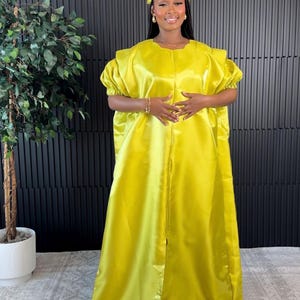 May include: A bright yellow satin dress with short sleeves and a matching headband. The dress has a loose, flowing design and a front slit. The background is a black wall with vertical lines, and a decorative tree is on the left.