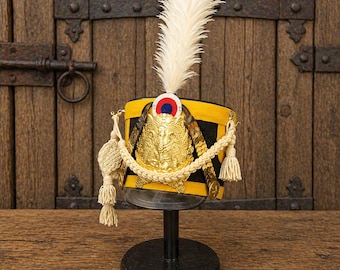 Puede incluir: Sombrero militar amarillo y negro con una gran pluma blanca, adornos dorados y una cuerda decorativa. El sombrero se exhibe sobre un soporte negro contra una puerta de madera. El sombrero tiene un emblema rojo, blanco y azul.
